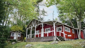 Une cabane rouge avec un porche et une petite construction adjacente, le tout entouré d’arbres.