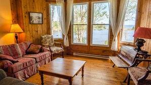 A cozy cabin interior with a wooden floor, a floral armchair, a red checkered bench, and a wooden table.