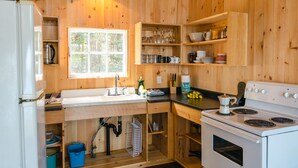 A compact kitchen with wooden walls, a window, a sink, a stove, and shelves with kitchenware.