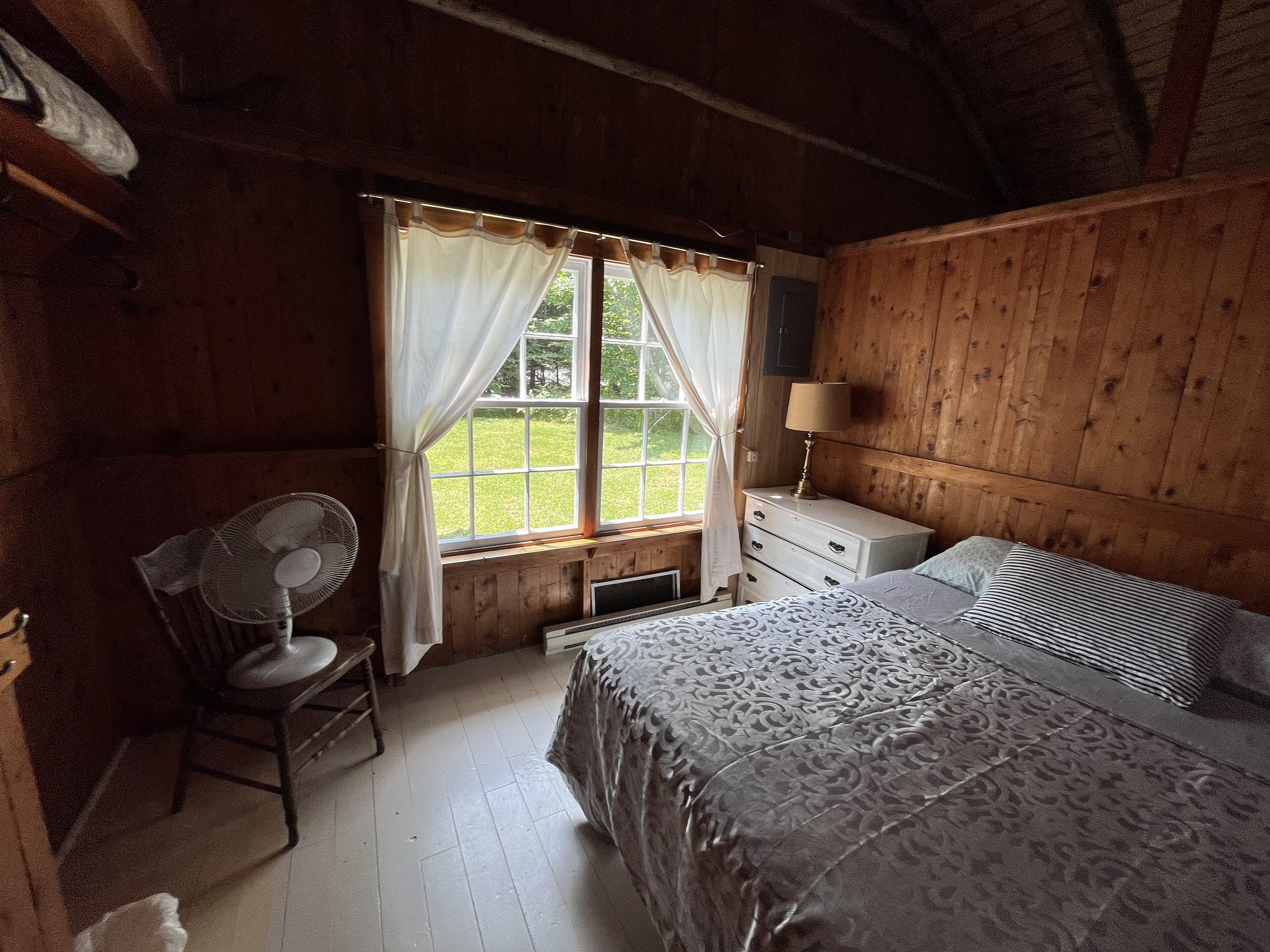 A wooden cabin bedroom with a bed, a chair, a desk, a lamp, and a window with curtains.