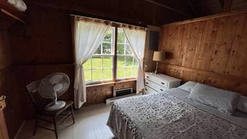 A wooden cabin bedroom with a bed, a chair, a desk, a lamp, and a window with curtains.