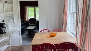 A dining area with a wooden table and red chairs, a white cabinet with shelves, and a window with striped curtains. - Beach Cottage - Pine Terrace - Hubbards Beach (Hubbards)
