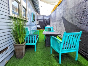 Un pequeño patio con una mesa y dos sillas, rodeado de césped artificial y una planta en maceta.