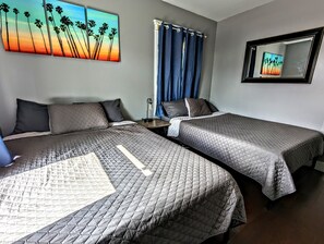 Una habitació d'hotel amb dos llits, una taula de nit, una làmpada, una finestra amb cortines blaves i un quadre de palmeres a la paret.