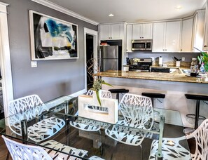 Una cocina moderna con gabinetes blancos, un refrigerador de acero inoxidable y un microondas. Hay un comedor con una mesa de vidrio y sillas blancas.