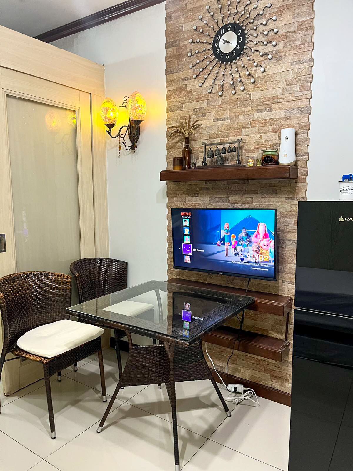 A living room with a glass-top dining table, wicker chairs, a flat-screen TV, a stone wall clock, and a fireplace mantel.