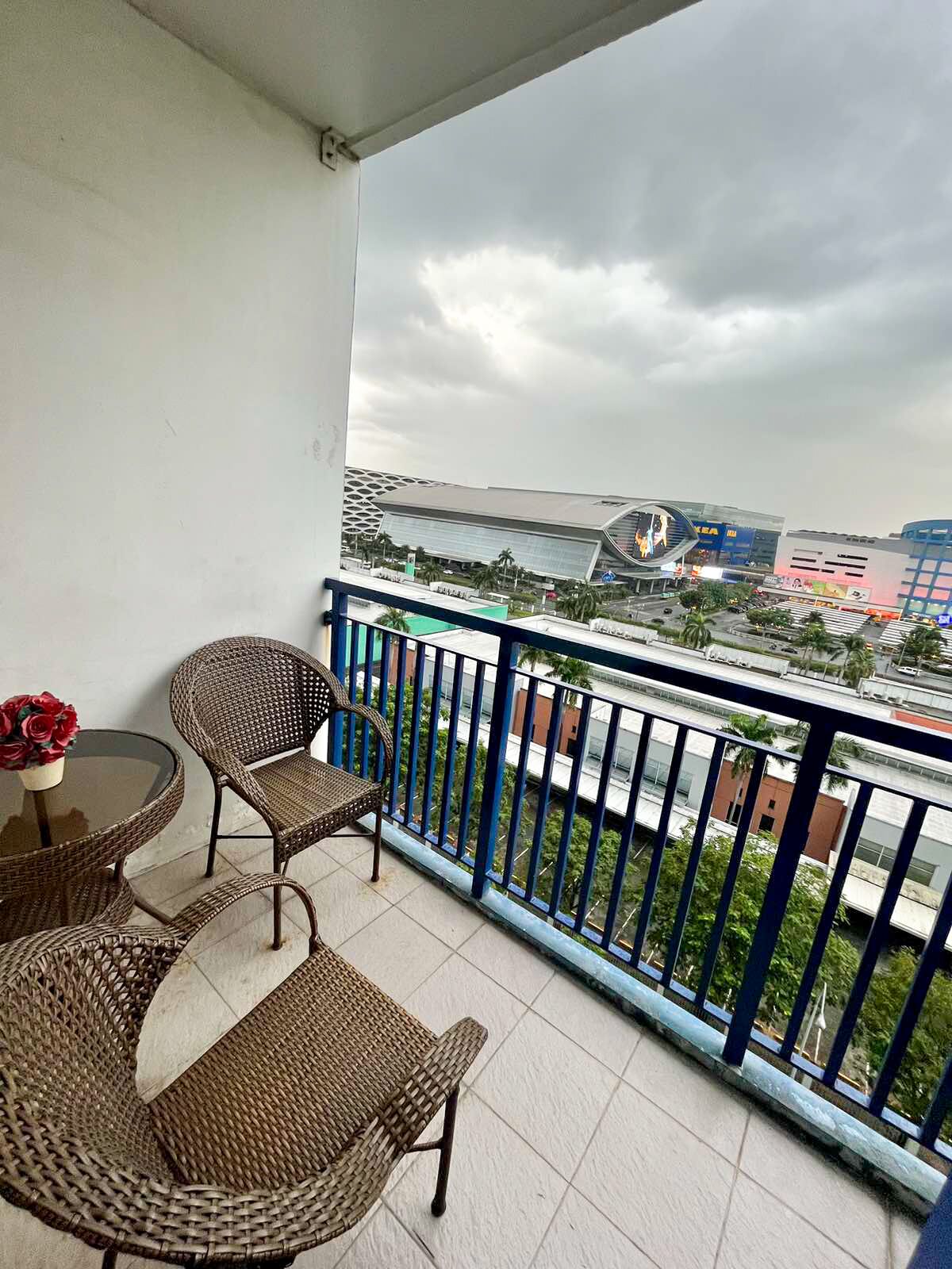 Een balkon met rieten meubels en een glazen tafel, met uitzicht op een stadsgezicht met moderne gebouwen en een grote constructie.