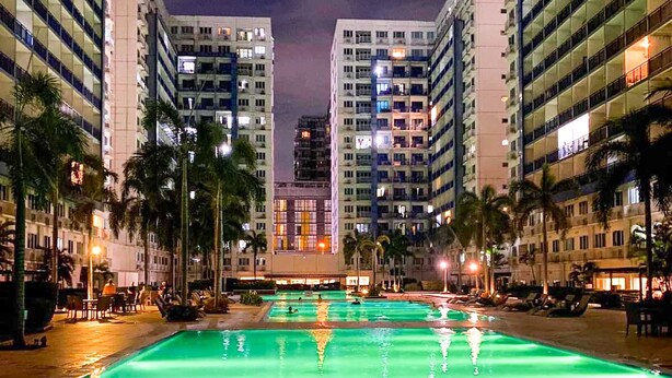 A nighttime view of a hotel with a central swimming pool, surrounded by high-rise buildings and palm trees. - Madison Place at Sea Residences powered by Cocotel (Pasay)
