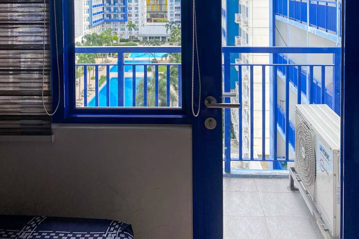 A room with a view of a high-rise building, a balcony with blue railings, and a pool.
