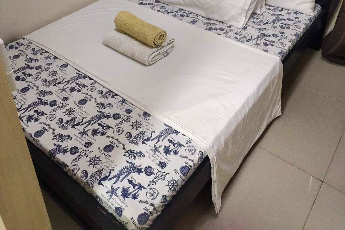 A single bed with a white and blue patterned bedspread, two white pillows, and a rolled-up towel on the bed.