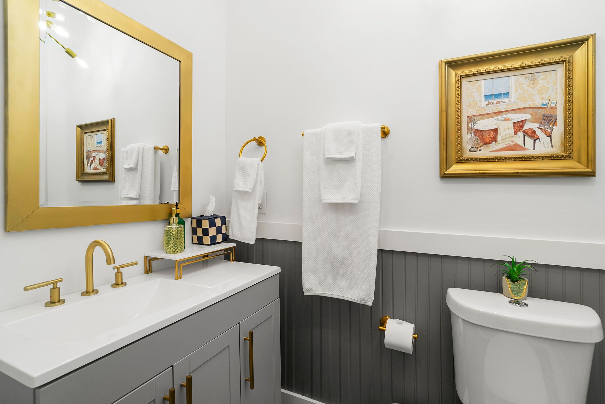A bathroom with a white sink, gold faucets, a gold-framed mirror, a toilet, and a framed picture on the wall.