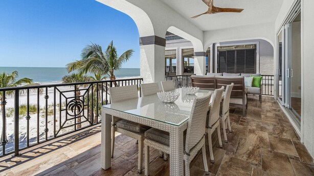 Ein Balkon mit einem Glastisch und Korbstühlen, der auf einen Strand mit Palmen und das Meer blickt.
