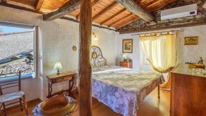 Ein rustikales Schlafzimmer mit Holzbalkendecke, einem Bett mit floralem Überwurf, einem Nachttisch mit Lampe und einem Fenster mit Blick auf eine Steinmauer.