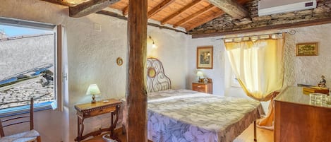 Ein rustikales Schlafzimmer mit Holzbalkendecke, einem Bett mit floralem Überwurf, einem Nachttisch mit Lampe und einem Fenster mit Blick auf eine Steinmauer.