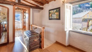 Ein Zimmer mit Holzbalkendecke, einem großen Fenster mit Blick auf Häuser, einem gerahmten Gemälde und einem Vintage-Koffer.