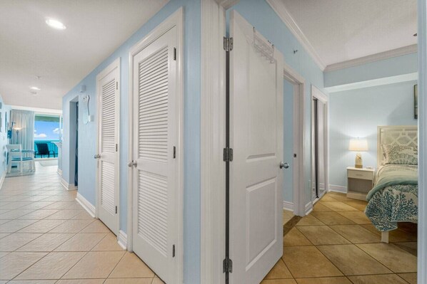 A hotel corridor with closed doors, a bed with a patterned bedspread, and a view of the ocean through an open door.