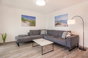 Un salon moderne avec un canapé d’angle gris, une table basse blanche et deux tableaux accrochés au mur représentant des paysages côtiers.