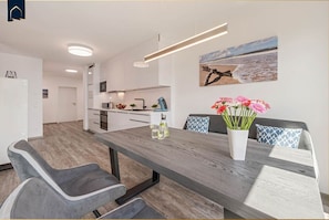 A modern kitchen with white cabinets, a wooden dining table, and a painting of a beach scene. - Ferienresidenz Am Haff - Neuerbautes Feriendomizil in Ruhiger Lage mit Blick auf das Stettiner Haff (Garz)