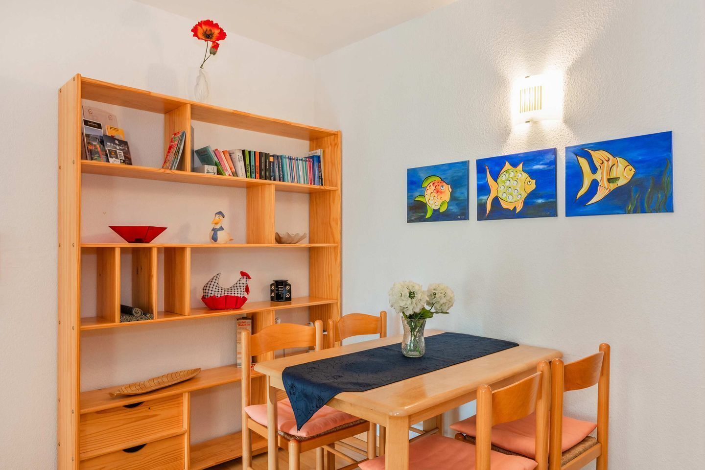 Una stanza con una libreria in legno, un tavolo da pranzo con sedie e tre quadri appesi al muro raffiguranti pesci.