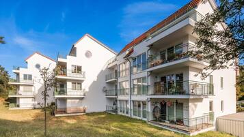 Un immeuble résidentiel à plusieurs étages, doté de balcons, d’un petit espace jardin et surplombant un ciel bleu dégagé.