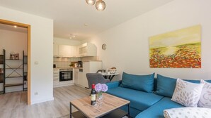 A modern living room with a blue sofa, a coffee table with a vase of flowers, a kitchen in the background, and a painting on the wall. - Ferienwohnung Kranich (Heringsdorf)