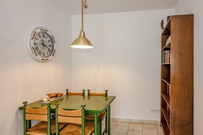 Ein Essbereich mit einem grünen Tisch, vier Stühlen, einer Wanduhr, einer Pendelleuchte und einem Holzregal.