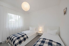 A hotel room with two single beds, a bedside table with a lamp, a hanging light fixture, and sheer curtains.