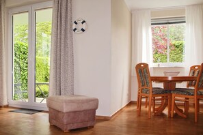 Una habitación con puerta de vidrio corredera que da a un jardín, una mesa de madera con sillas y un pequeño otoman.