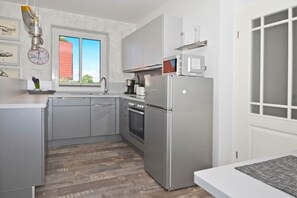 Et moderne køkken med lysegrå skabe, et stålskab, en indbygget ovn og et vindue med udsigt til en rød bygning.