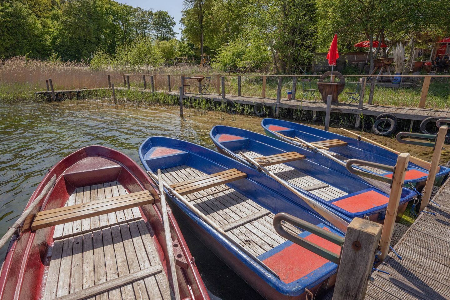 A row of wooden rowboats docked at a pier with a wooden walkway and a red umbrella in the background.
