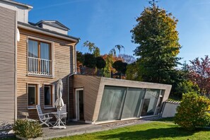 Ein modernes, zweistöckiges Haus mit Balkon, einer Terrasse mit Gartenmöbeln und einem großen Baum im Hintergrund.