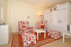 Ein Wohnzimmer mit zwei floralen Ohrensesseln, einem kleinen weißen Tisch, einer Couch, einem Fernseher und einem Bücherregal.