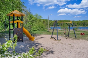 Un parque infantil con tobogán y columpios, rodeado de árboles y un cuerpo de agua.