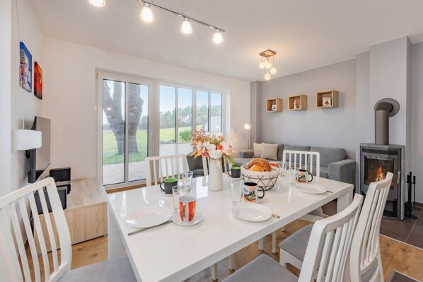 Un comedor moderno con una mesa blanca dispuesta para cuatro personas, una chimenea y vistas al exterior.