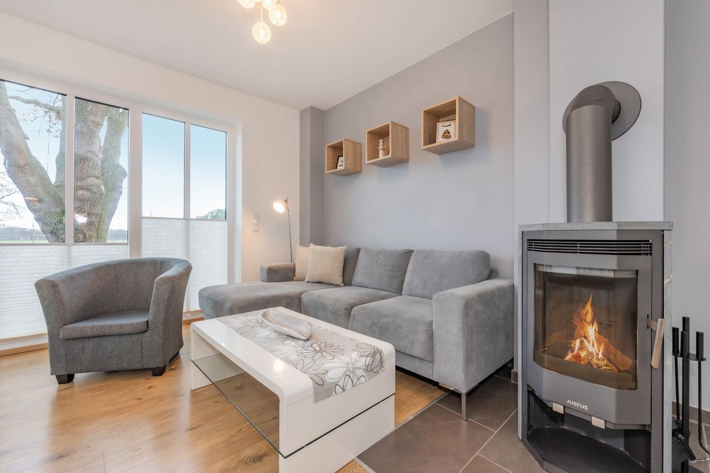 A modern living room with a grey sofa, a grey armchair, a glass coffee table, and a wood-burning stove.