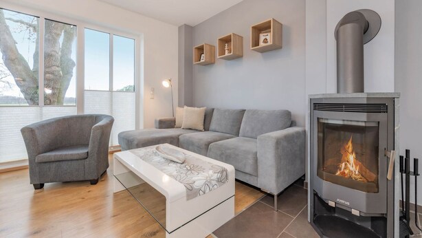 Ein modernes Wohnzimmer mit einem grauen Sofa, einem grauen Sessel, einem Glastisch und einem Kaminofen.