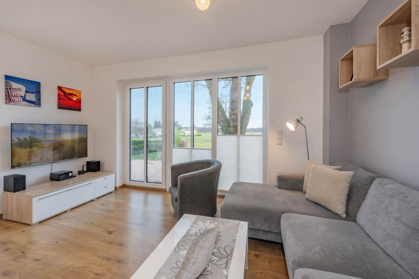 A modern living room with a flat-screen TV, a grey sofa, a wooden coffee table, and a large sliding glass door leading to a garden.