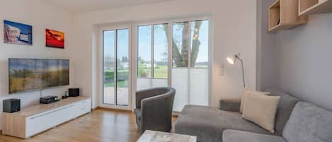 A modern living room with a flat-screen TV, a grey sofa, a wooden coffee table, and a large sliding glass door leading to a garden.