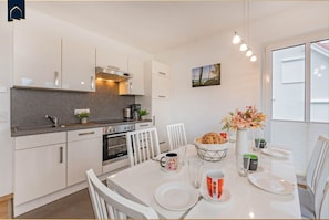 A modern kitchen with white cabinets, a built-in oven, and a dining area with a white table set for two. - DHH Anemone - Gemütliche Doppelhaushälfte für bis zu 6 Personen, mit Kamin und Terrasse (Korswandt)