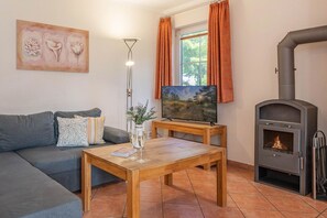 Un salon avec un canapé, une table basse sur laquelle se trouvent un livre et des lunettes, un téléviseur posé sur un meuble, des rideaux orange et un poêle à bois.