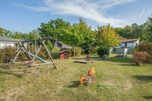 Ein Spielplatz mit Schaukel, Rutsche, einem kleinen roten Spielhäuschen und einer Wippe in einem grünen Garten mit Bäumen und Häusern im Hintergrund.