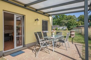 Eine überdachte Terrasse mit Tisch und Stühlen, eine Glastür zu einem Innenraum und Blick auf einen umzäunten Garten.