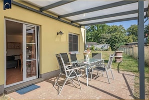 Une terrasse couverte avec une table et des chaises, une porte-fenêtre coulissante donnant sur une pièce intérieure et une vue sur un quartier résidentiel.