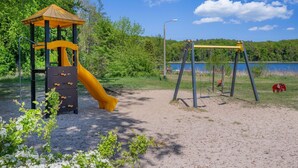 A playground with a slide and swings, surrounded by trees and a lake. - Ferienwohnung Waldhaus 2.2 - Gemütlicher Ferienbungalow in Ruhiger Waldrandlage, 800 m vom (Korswandt)