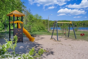 Un parque infantil con tobogán y columpios, rodeado de árboles y un lago.