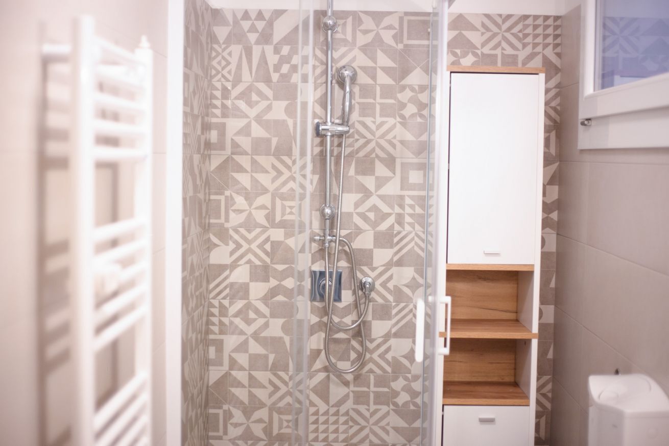 Eine Dusche mit geometrischem Fliesenmuster, einem weißen Schrank und einem Holzregal.