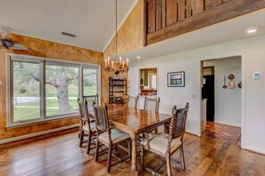 A spacious dining area with a large wooden table and chairs, a chandelier, and a view of a tree through large windows.