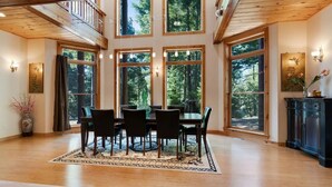 Une salle à manger spacieuse avec une table et des chaises en bois, de grandes fenêtres donnant sur la forêt et un vase décoratif posé sur le sol.
