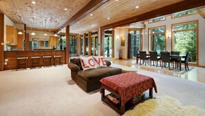Un vaste espace de vie doté d’un plafond en bois, d’un canapé agrémenté d’un coussin orné du mot « AMOUR », d’une table à manger avec des chaises, et de grandes fenêtres offrant une vue sur l’extérieur.