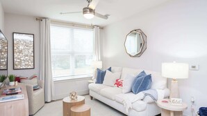 Una sala de estar moderna con un sofá blanco, mesas auxiliares de madera redondas, un ventilador de techo y un ventanal con persianas.